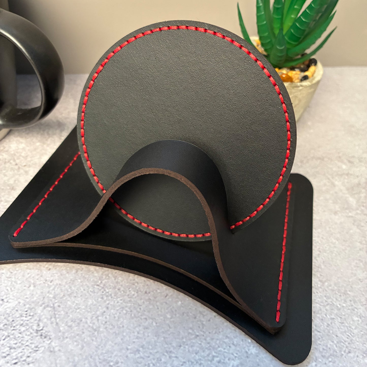 Round black leather coasters with red stitching in a matching leather coaster holder on a textured surface with a plant in the background