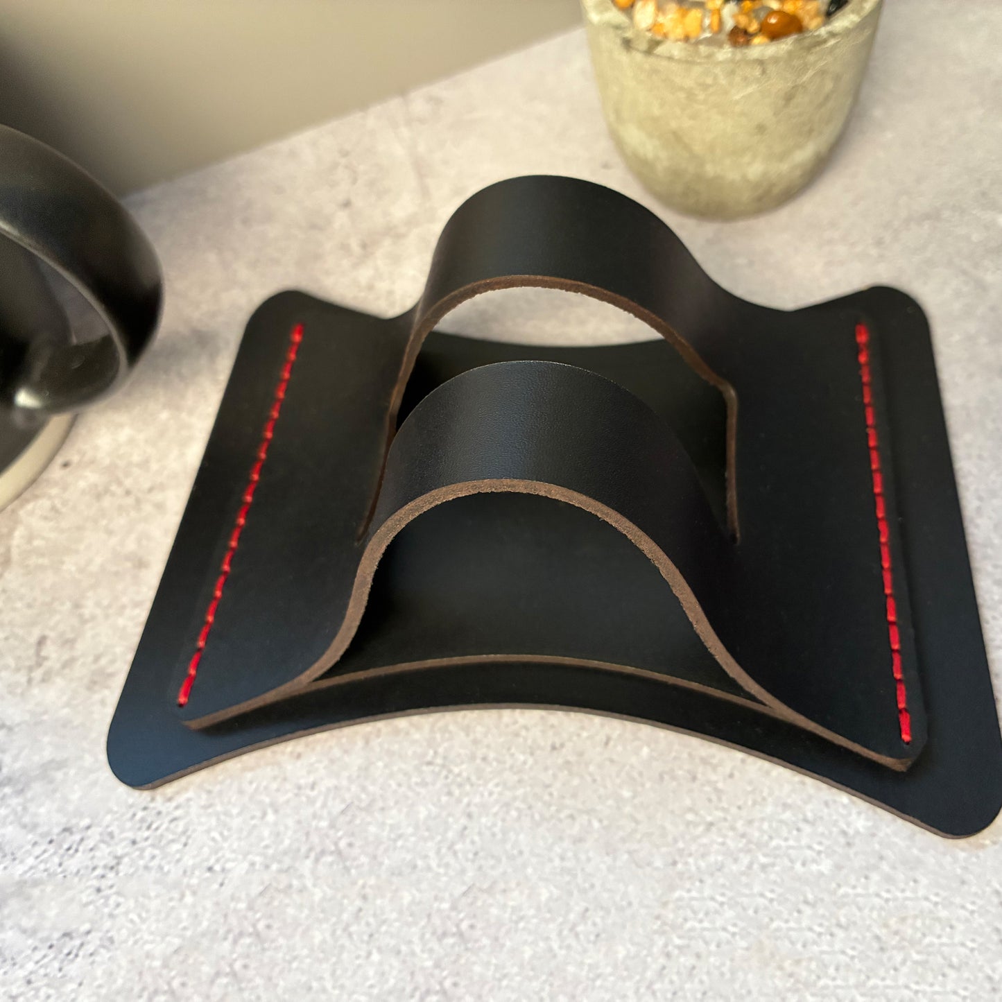Black leather coaster holder with red stitching on a textured surface.