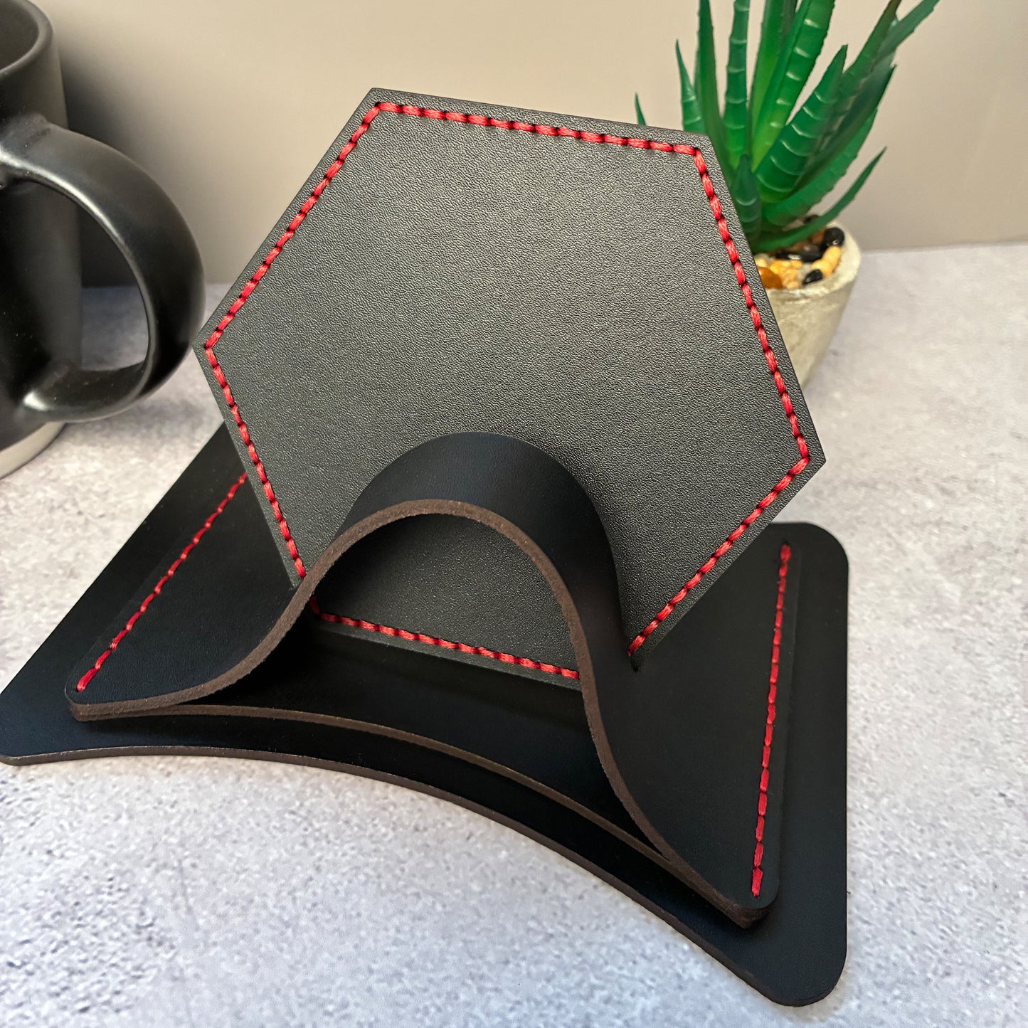 Hexagonal black leather coasters with red stitching in a matching leather coaster holder on a textured surface with a plant in the background