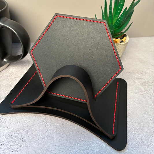 Hexagonal black leather coasters with red stitching in a matching leather coaster holder on a textured surface with a plant in the background