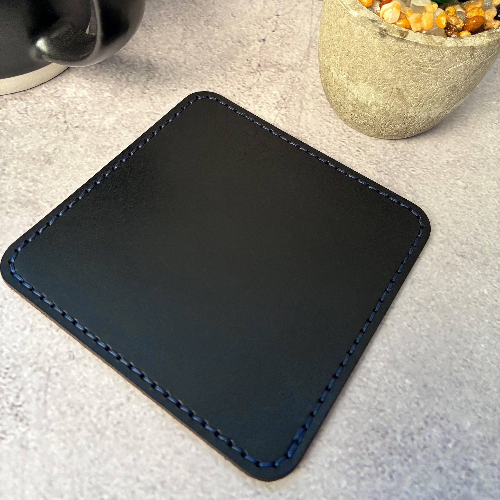 Black vegetable tanned leather square coaster with dark blue stitching on a light grey surface