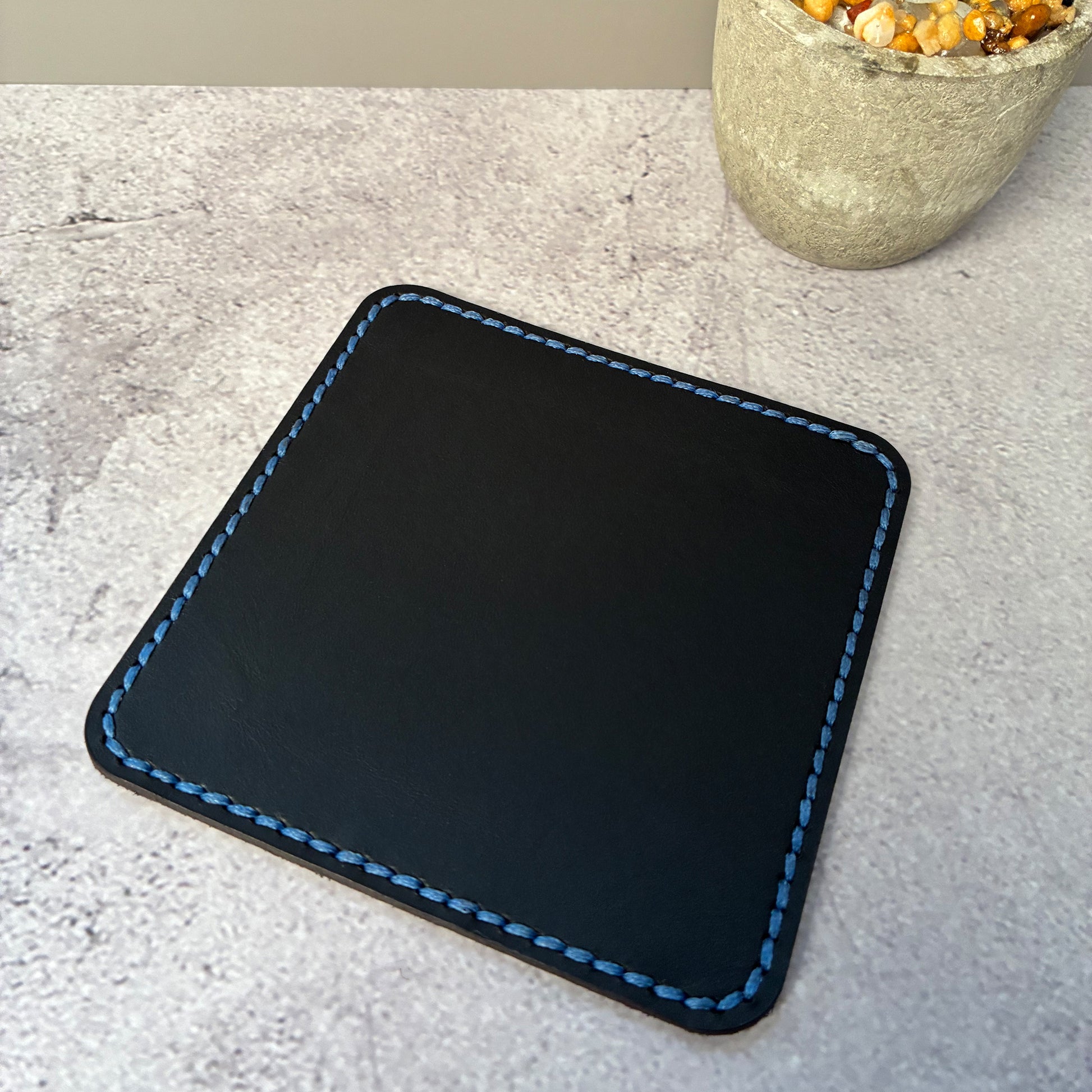 Black vegetable tanned leather square coaster with light blue stitching on a light grey surface