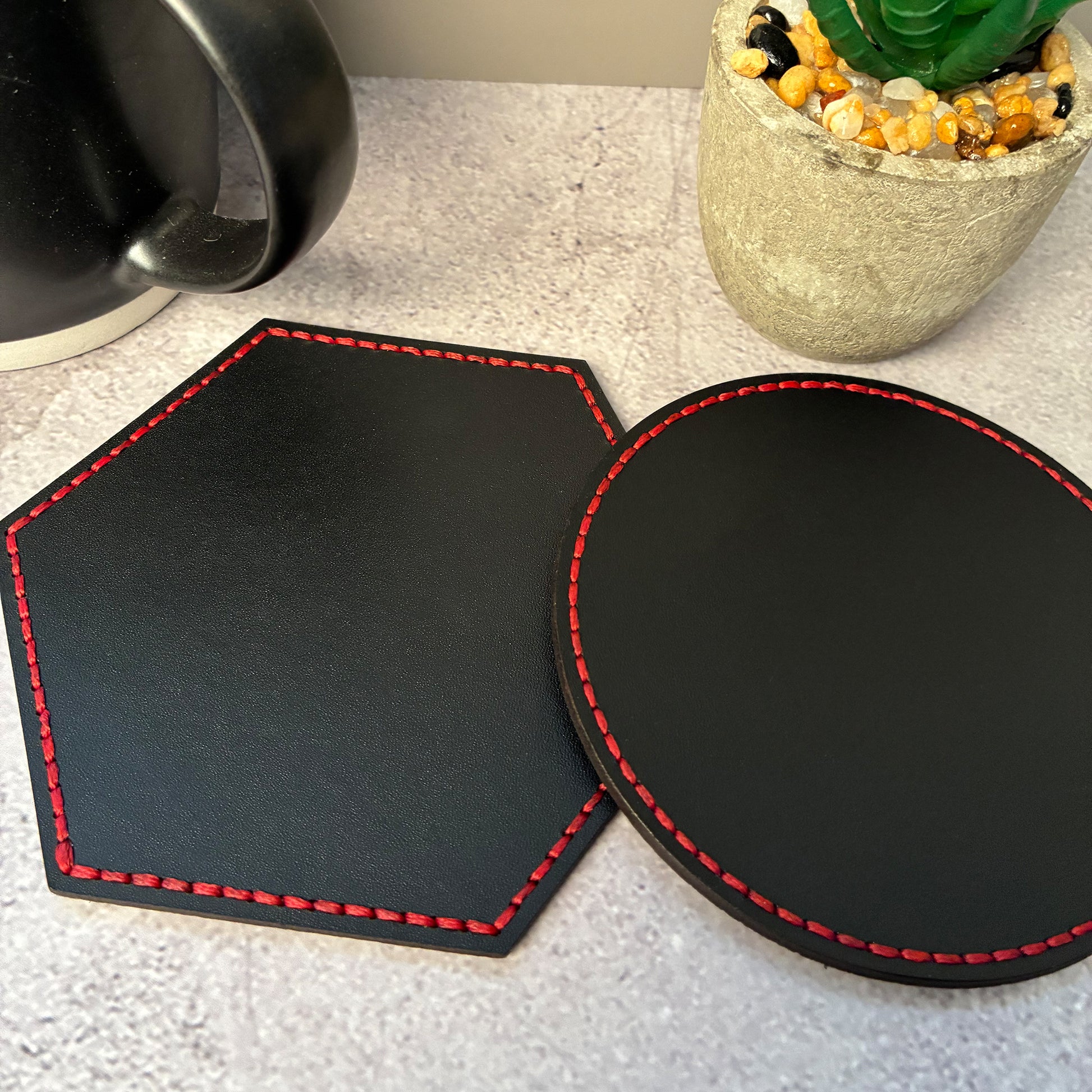 Black vegetable tanned leather coasters, round and hexagonal with red stitching on a light grey surface