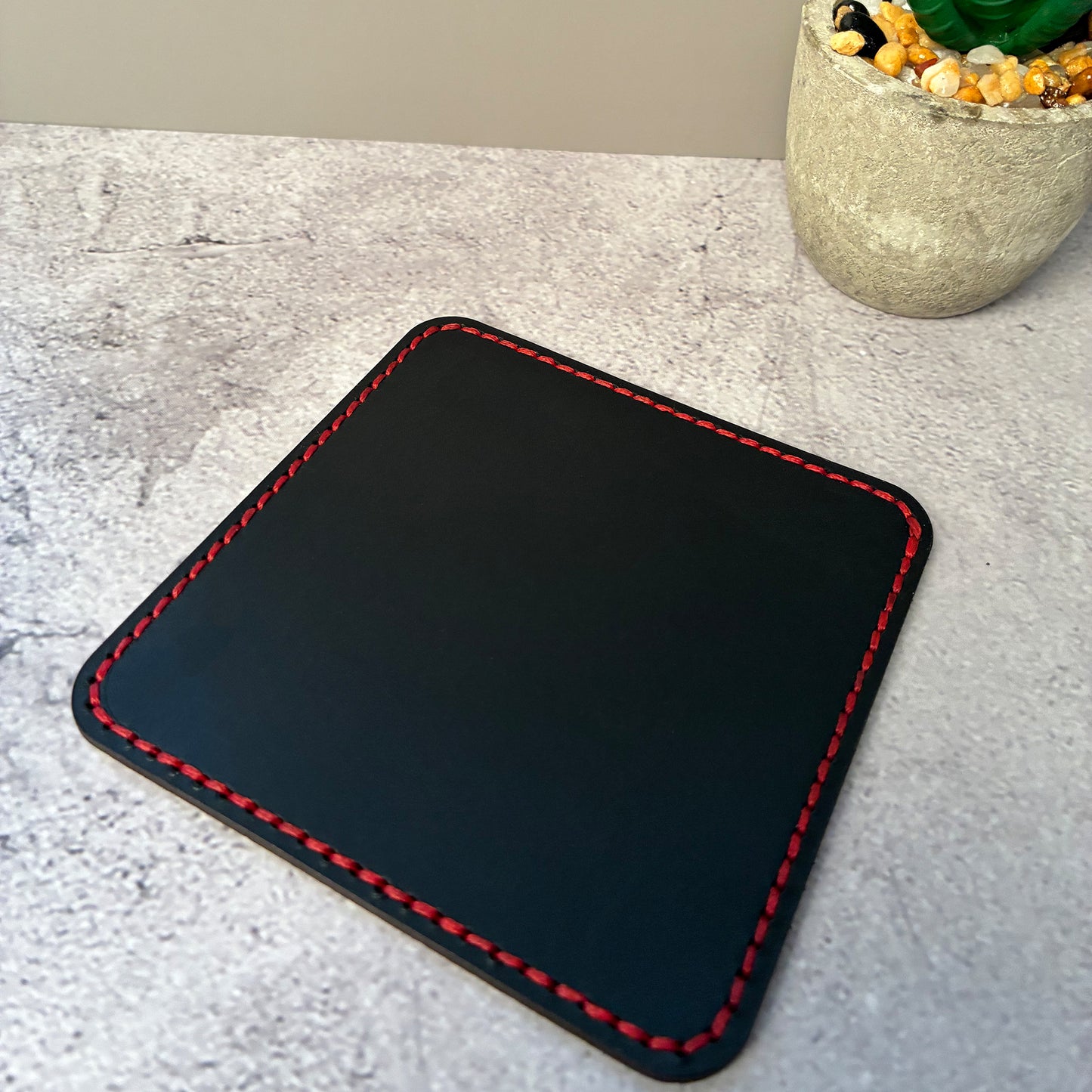 Black vegetable tanned leather square coaster with red stitching on a light grey surface