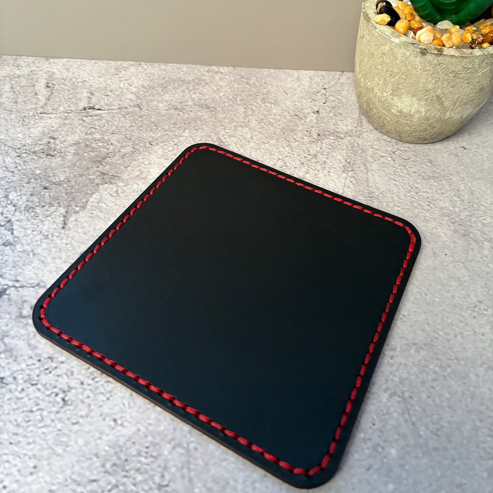 Black vegetable tanned leather square coaster with red stitching on a light grey surface