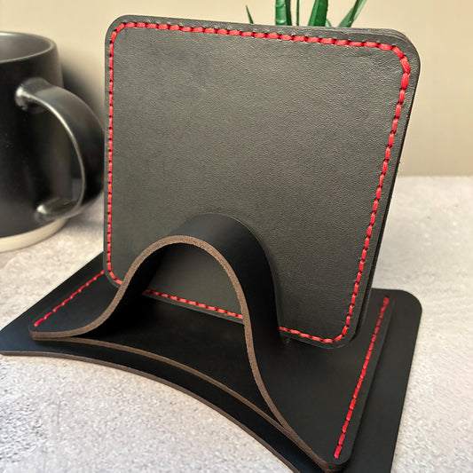 Set of black leather coasters with red stitching in a coaster holder, next to a mug and plant.