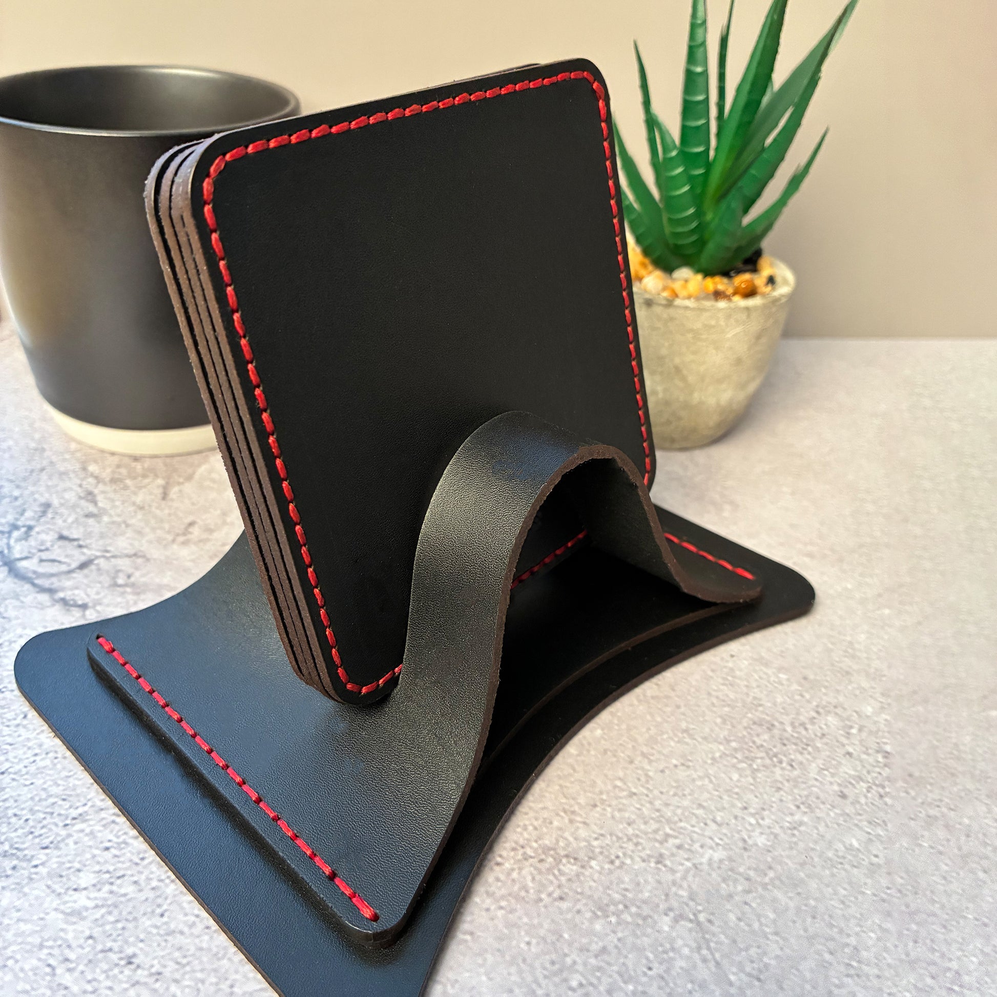 Set of black leather coasters with red stitching in a coaster holder, next to a mug and plant.