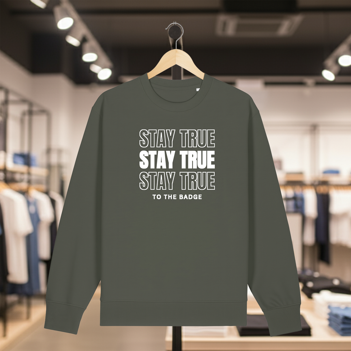 Khaki sweatshirt with text STAY TRUE TO THE BADGE on a hanger in a store setting